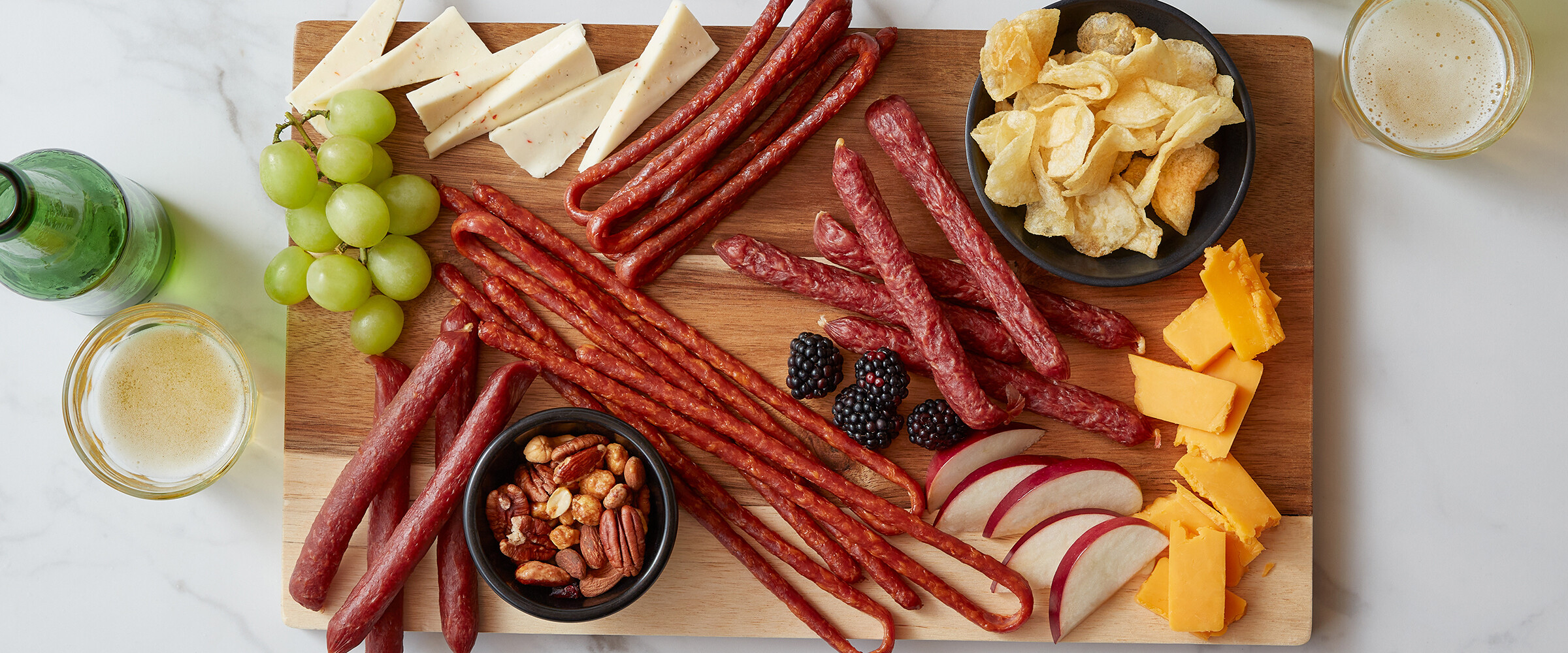 Salami whips and sticks on a board with grapes, cheese and chips