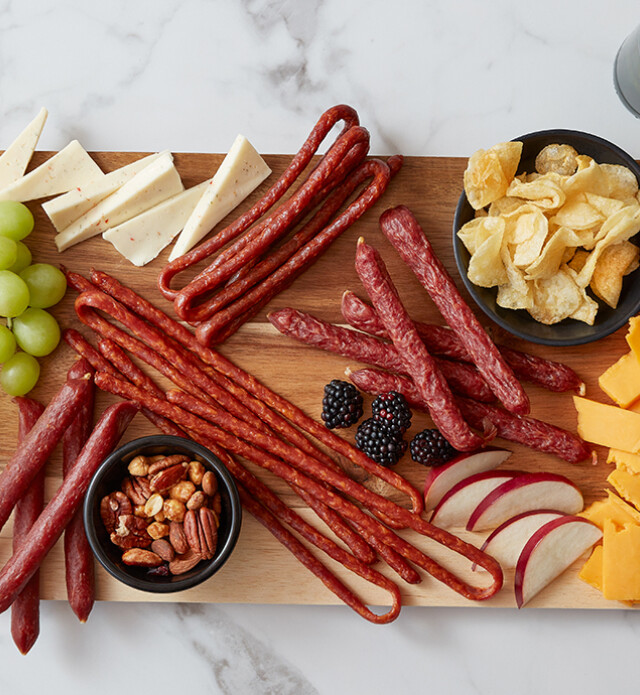 Salami whips and sticks on a board with grapes, cheese and chips